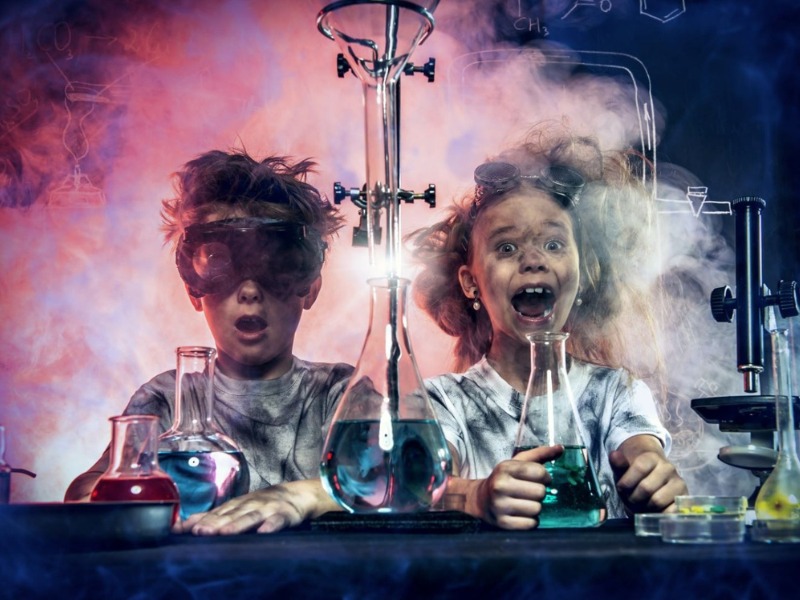 Two children with excited expressions conducting a science experiment that has produced smoke and coloured liquids. They are wearing safety goggles and appear messy from the experiment.