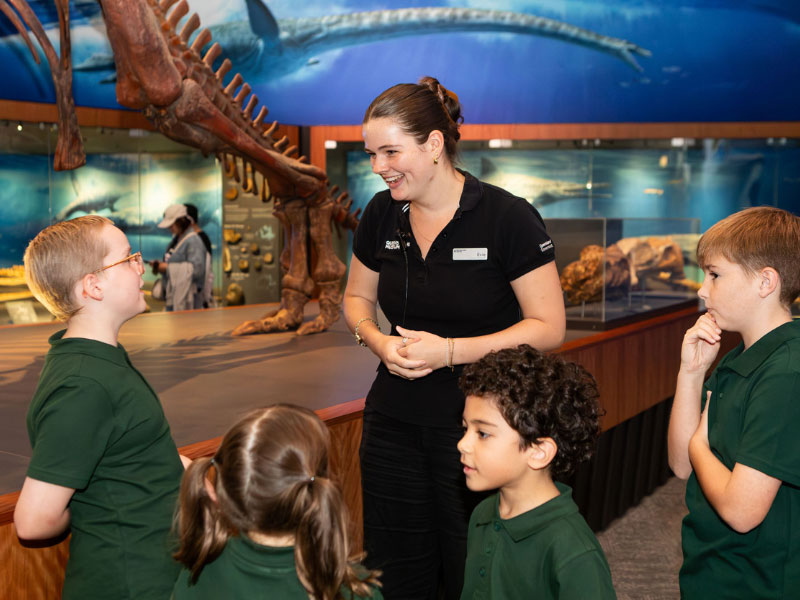 A museum educator speaking to a group of primary school students inside the Dinosaurs Unearthed exhibition at Queensland Museum Kurilpa